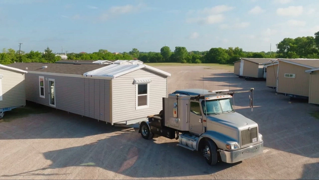 Modular homes parked beside a tractor-trailer at a transport yard.
